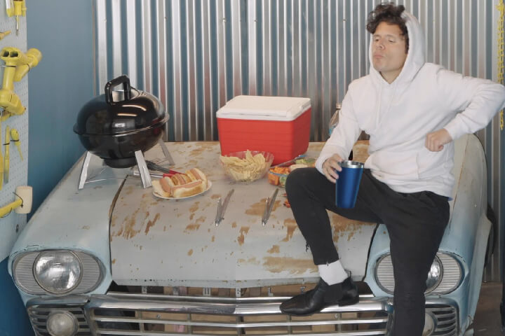 Man in white hoodie and black pants sits on the front of a car in a clearly-built set. There is a small grill, a plate of hotdogs, cooler, and chips also on the hood of the car.