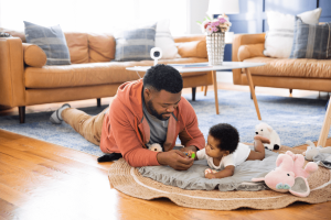 Man and baby play on the floor of a living room. Baby monitor in the background